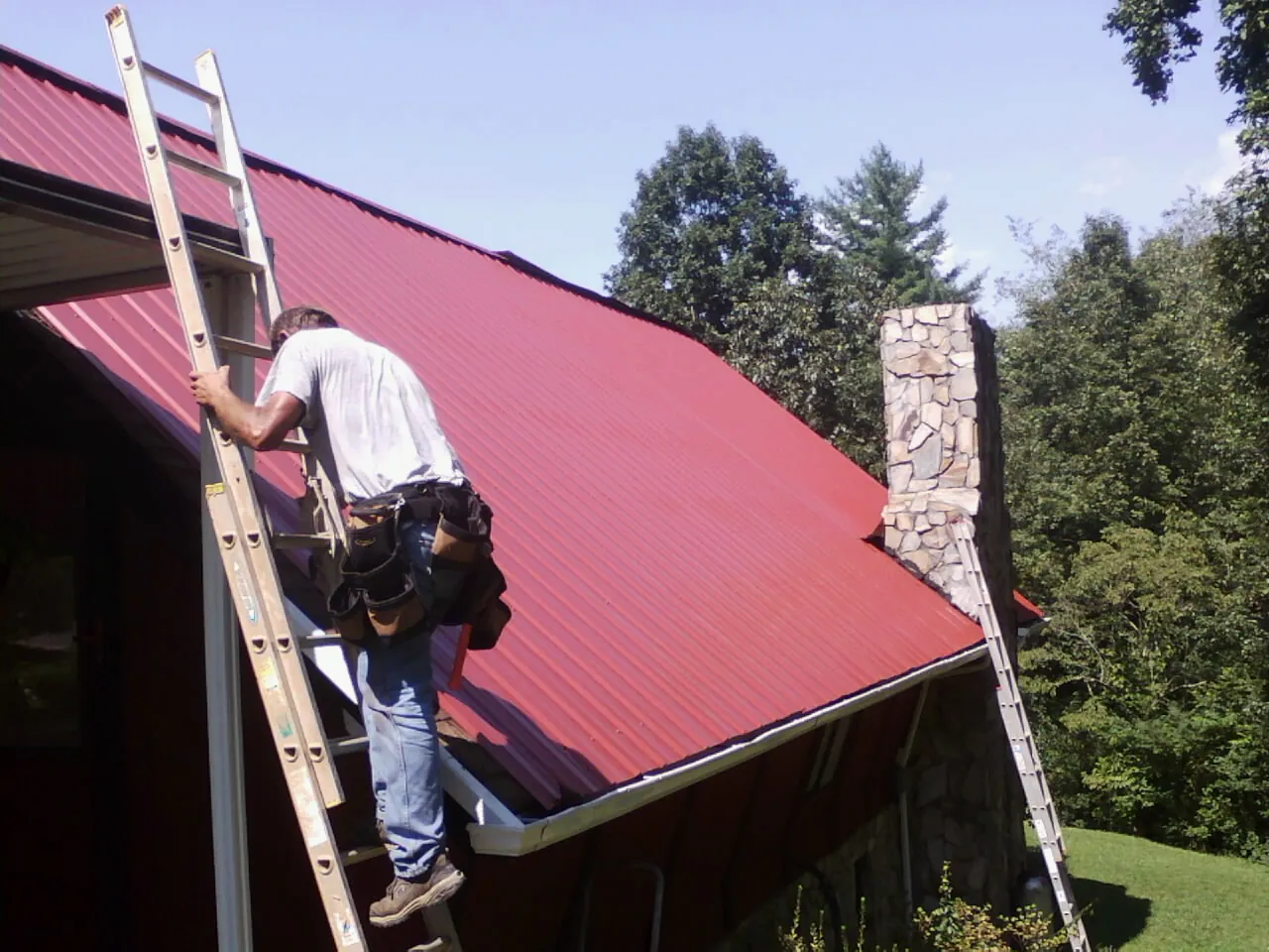 Roofer on a ladder beside a red metal roof and stone chimney — Soffit & Fascia Repair by Roofing Brothers in Hamler